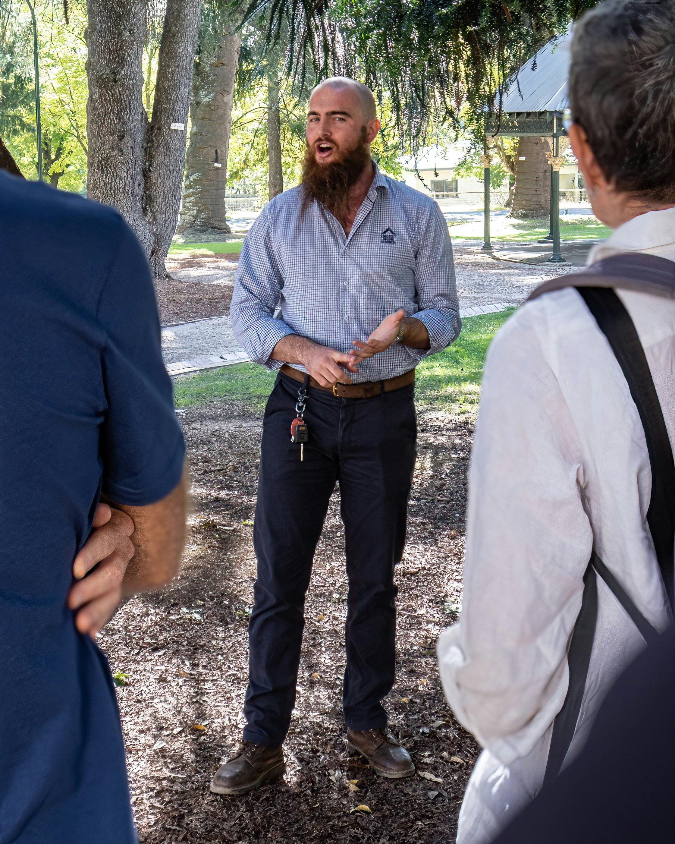 Tour guide with a long beard talking to two people in gardens with large trees in the background