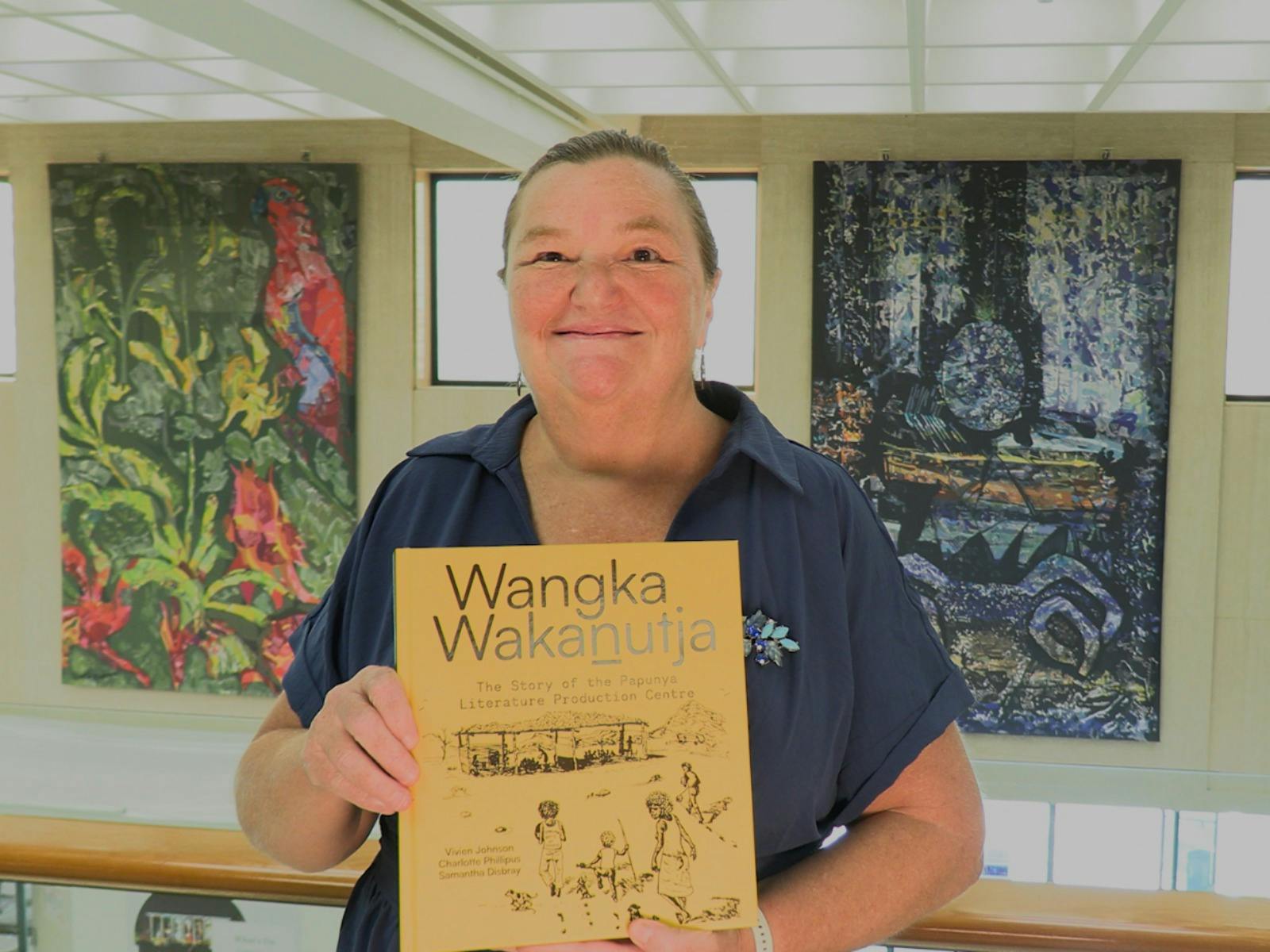 A woman holding a book with a yellow cover, standing in front of some wall tapestries