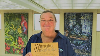A woman holding a book with a yellow cover, standing in front of some wall tapestries