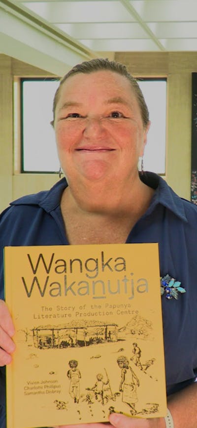 A woman holding a book with a yellow cover, standing in front of some wall tapestries