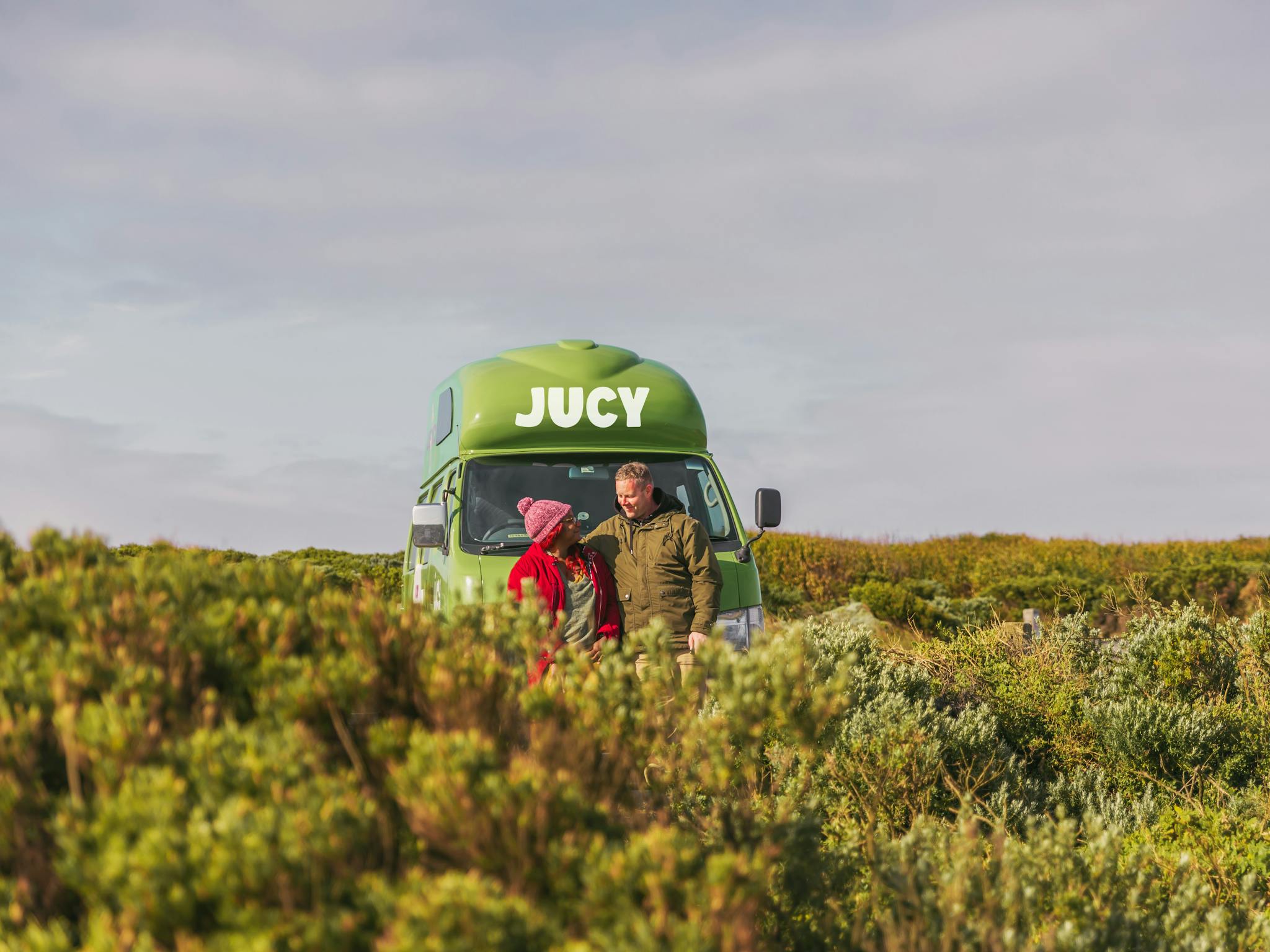 A couple standing in a farm in front of a JUCY green campervan in australia