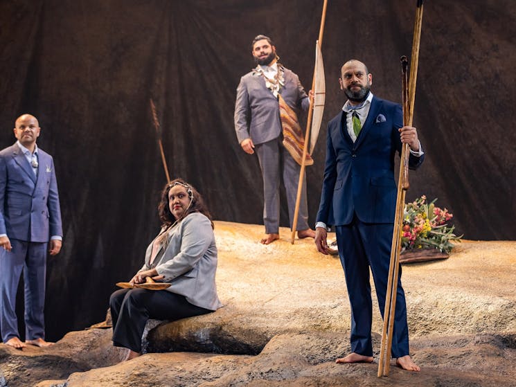 Image of four actors as First Nations Elders sitting and standing on a rock, all wearing suits