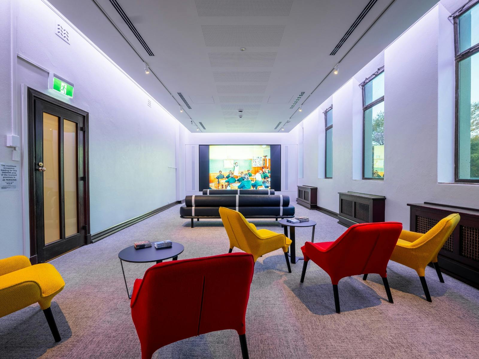 A light filled space with yellow and red chairs and a large screen playing documentaries