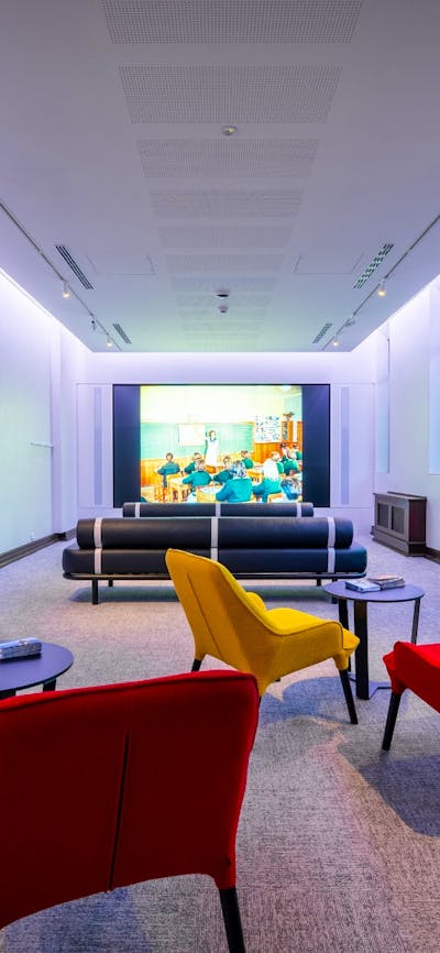 A light filled space with yellow and red chairs and a large screen playing documentaries