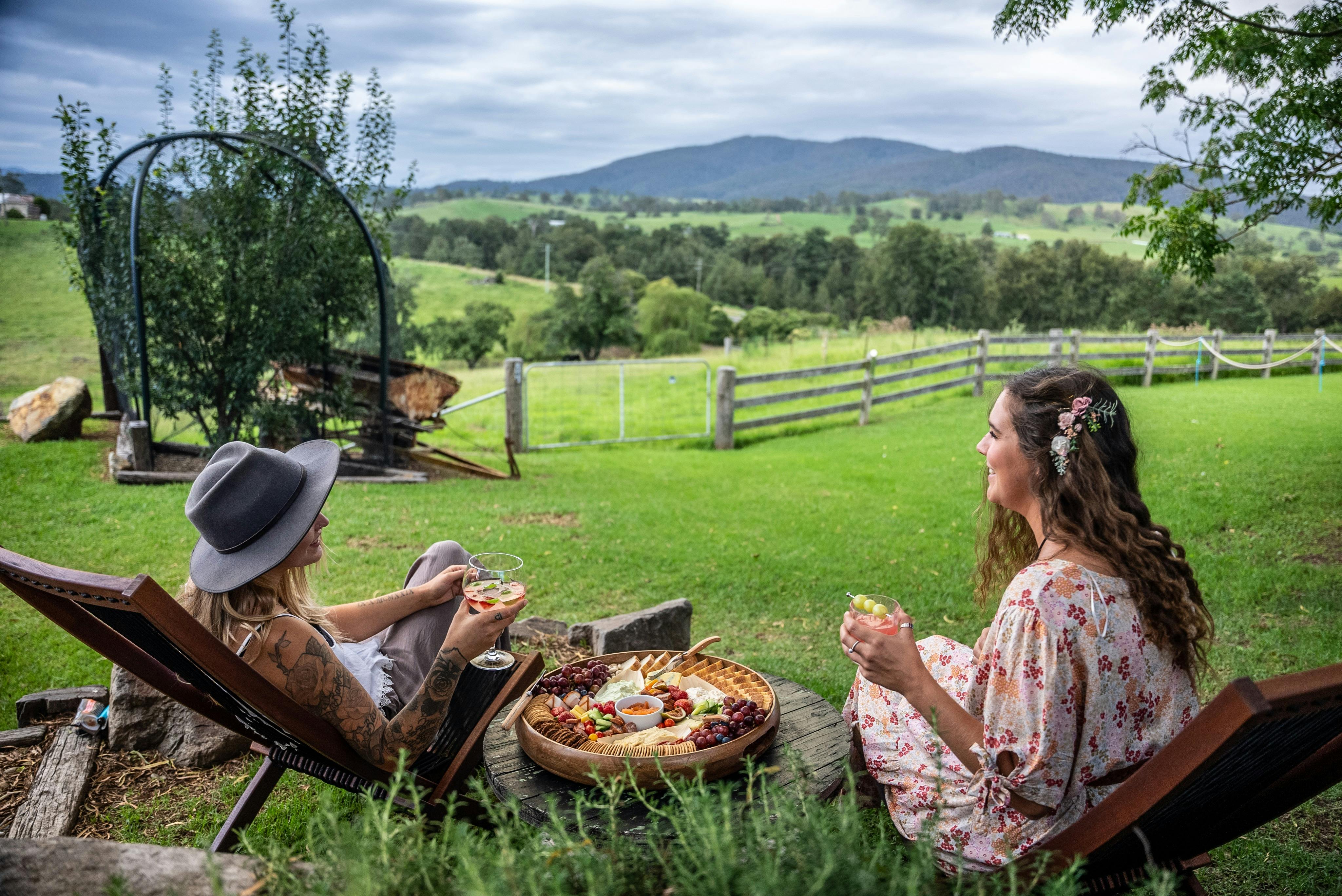 Zwei Frauen genießen in einem Garten Getränke und ein Picknick mit Blick auf sanfte grüne Hügel.