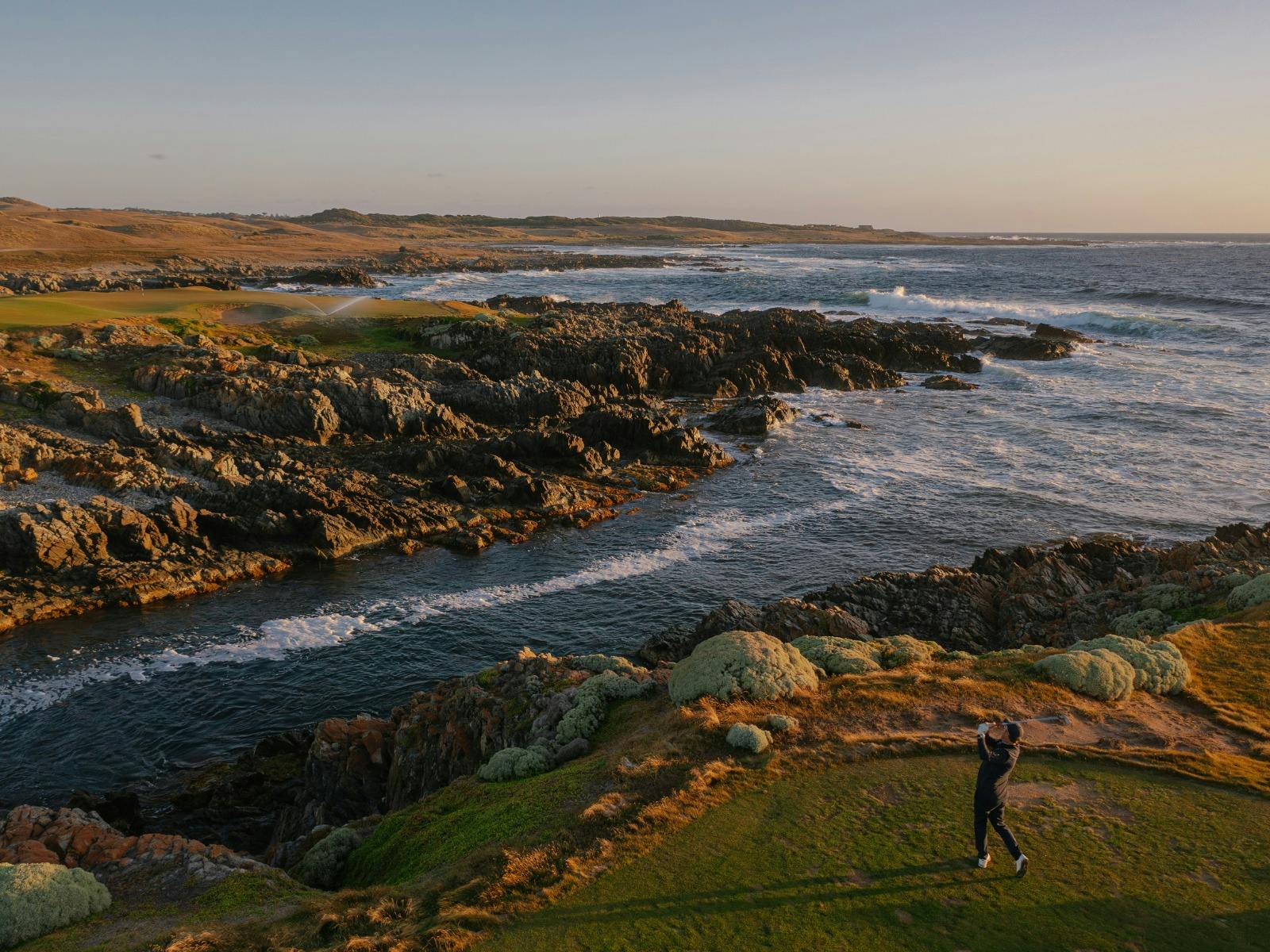 Golfer teeing off beside rugged coastline at Ocean Dunes during golden hour