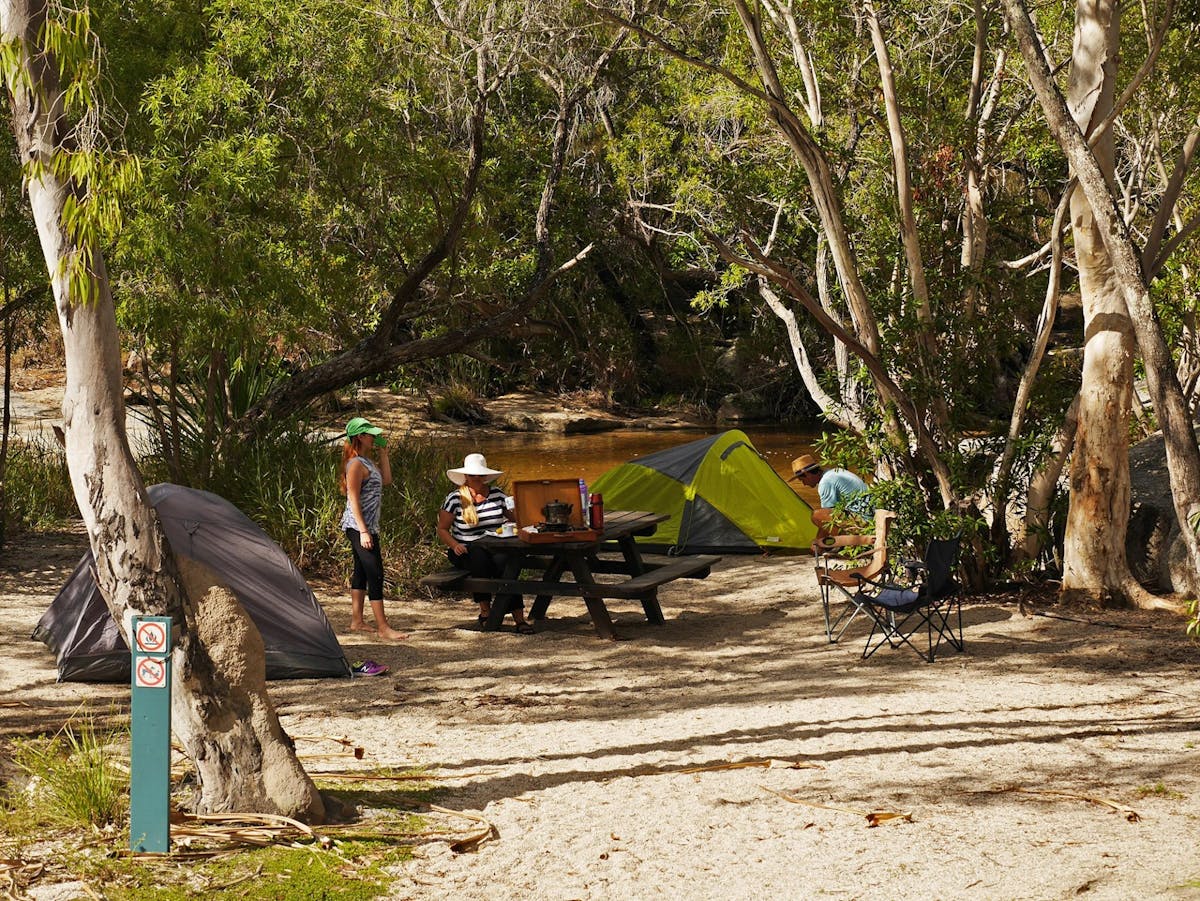 Two tents set up beside a creek with a picnic table and three people between the tents.