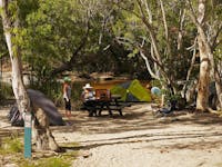 Two tents set up beside a creek with a picnic table and three people between the tents.