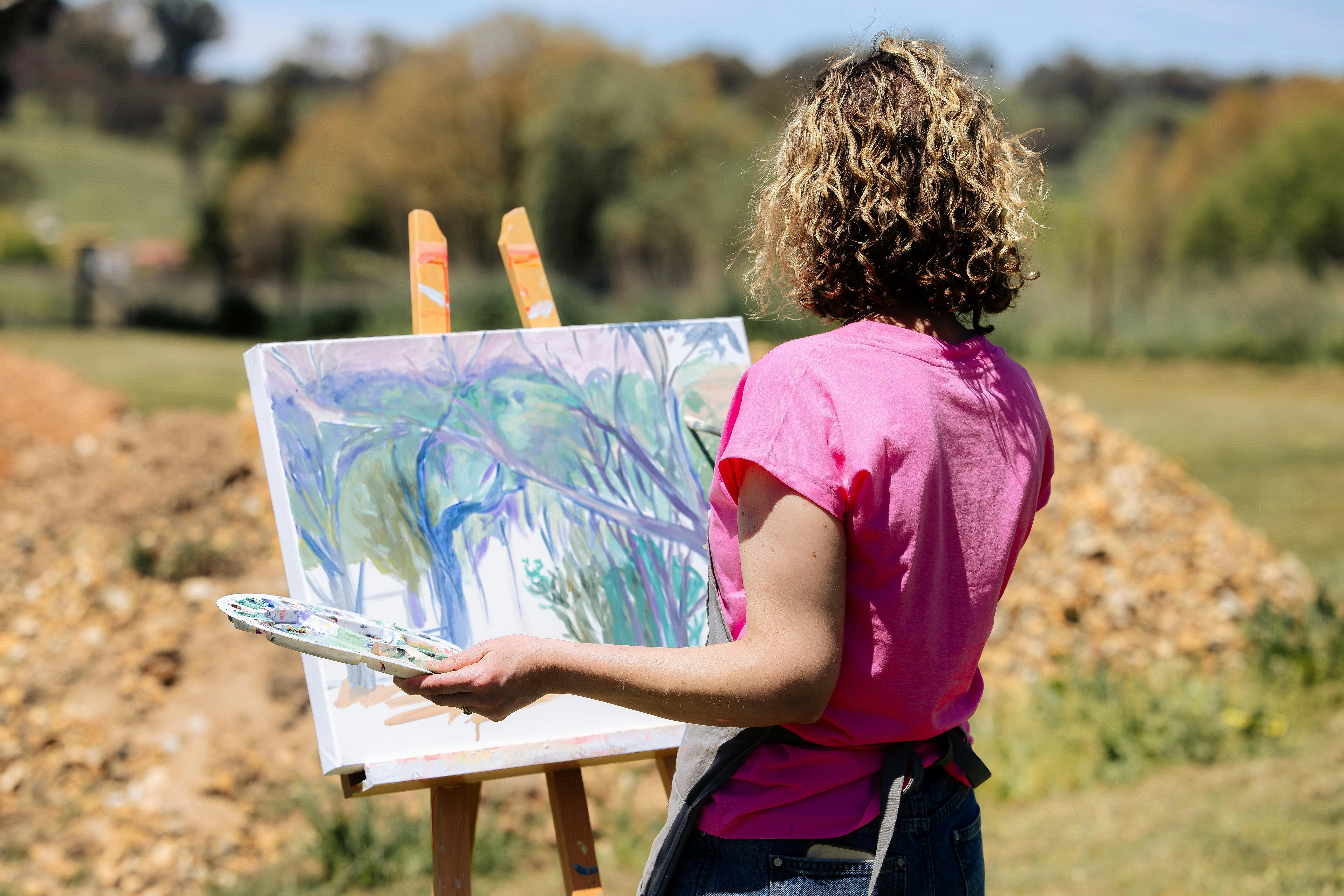 A woman in a pink top is viewed from behind painting on an easel in a rural setting