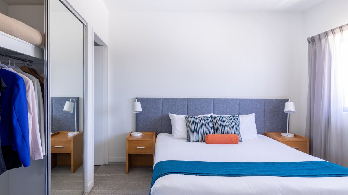 Bed with wooden bedside tables, a blue runner on the end and clothes in a wardrobe.