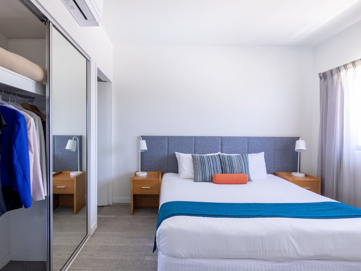 Bed with wooden bedside tables, a blue runner on the end and clothes in a wardrobe.