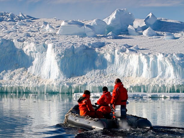 Three people in a small inflatable boat looking at the large ice formation in front of them.