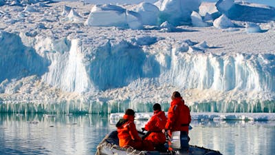 Three people in a small inflatable boat looking at the large ice formation in front of them.