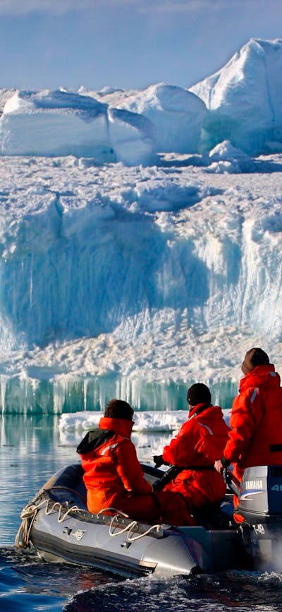 Three people in a small inflatable boat looking at the large ice formation in front of them.