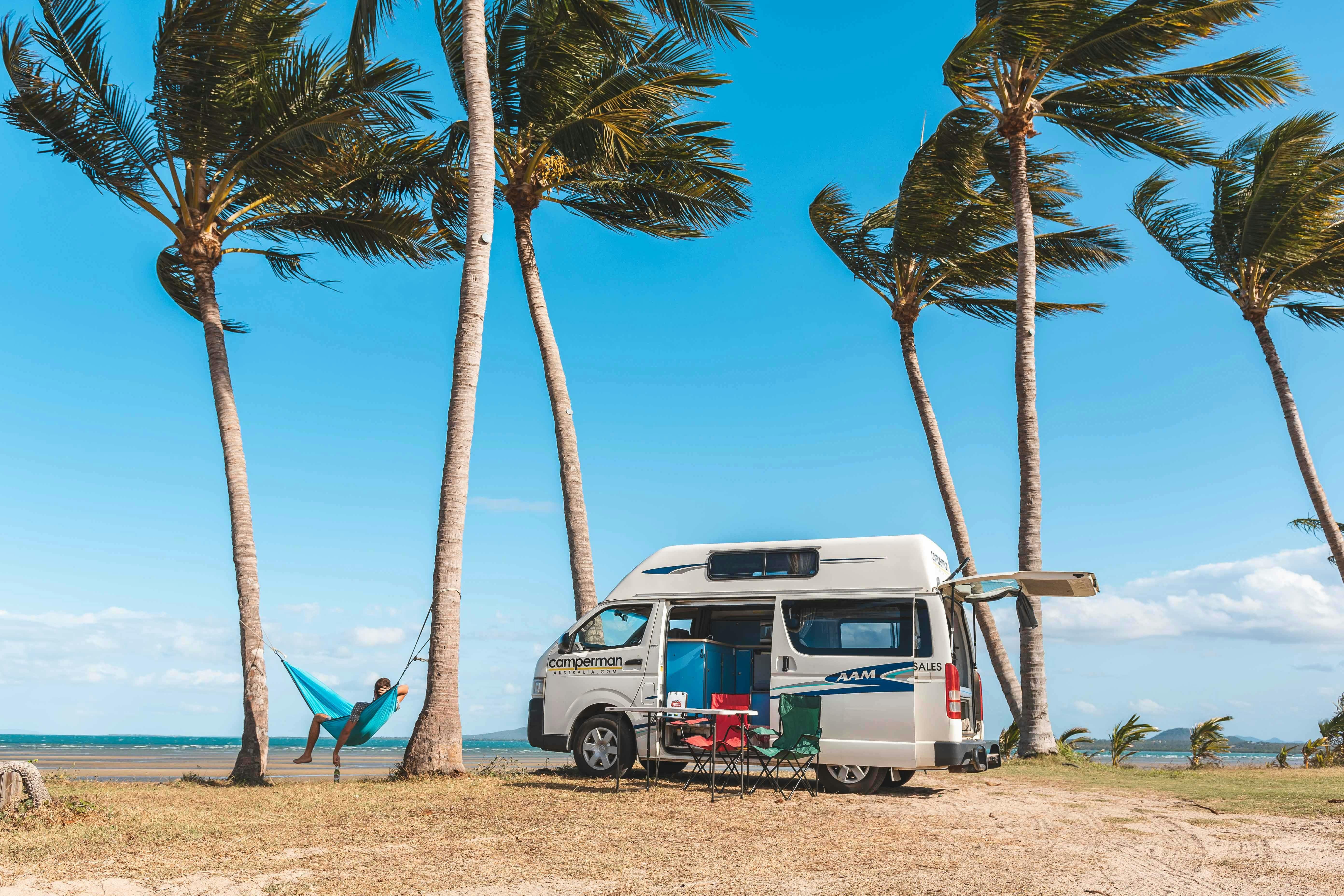 Person relaxing in a hammock on the beach under the palm trees, next to a campervan