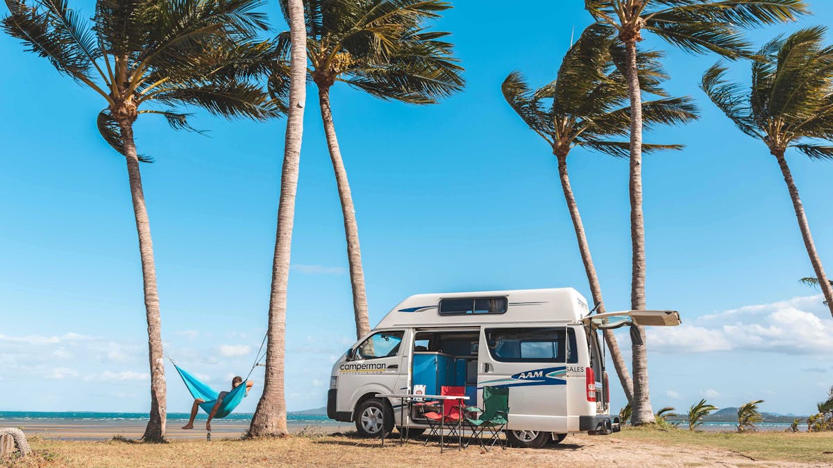 Person relaxing in a hammock on the beach under the palm trees, next to a campervan