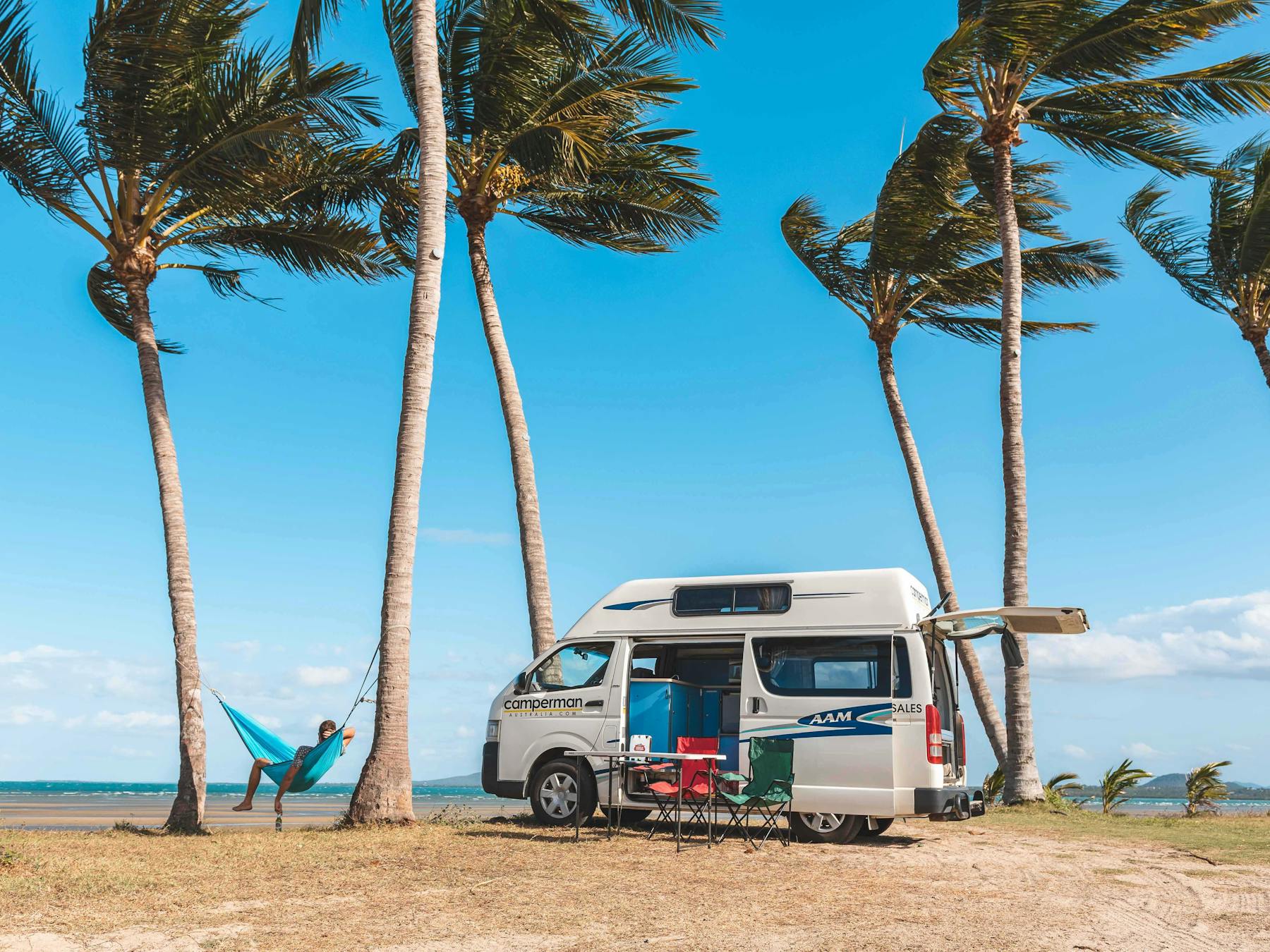 Person relaxing in a hammock on the beach under the palm trees, next to a campervan