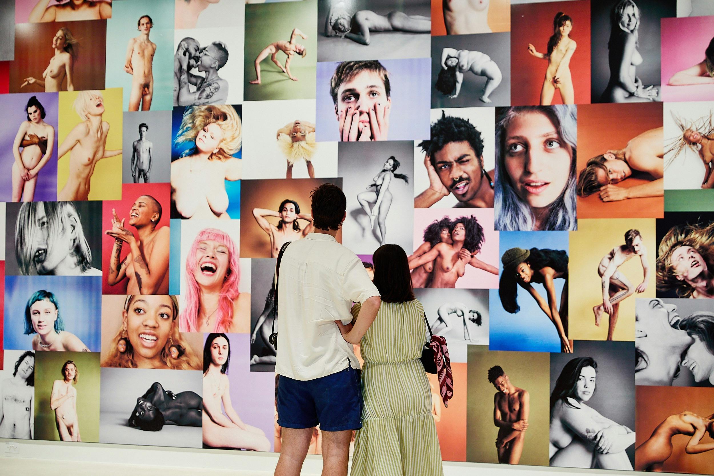 A man and woman view dozens of photos of nude models wallpapered to a gallery wall.