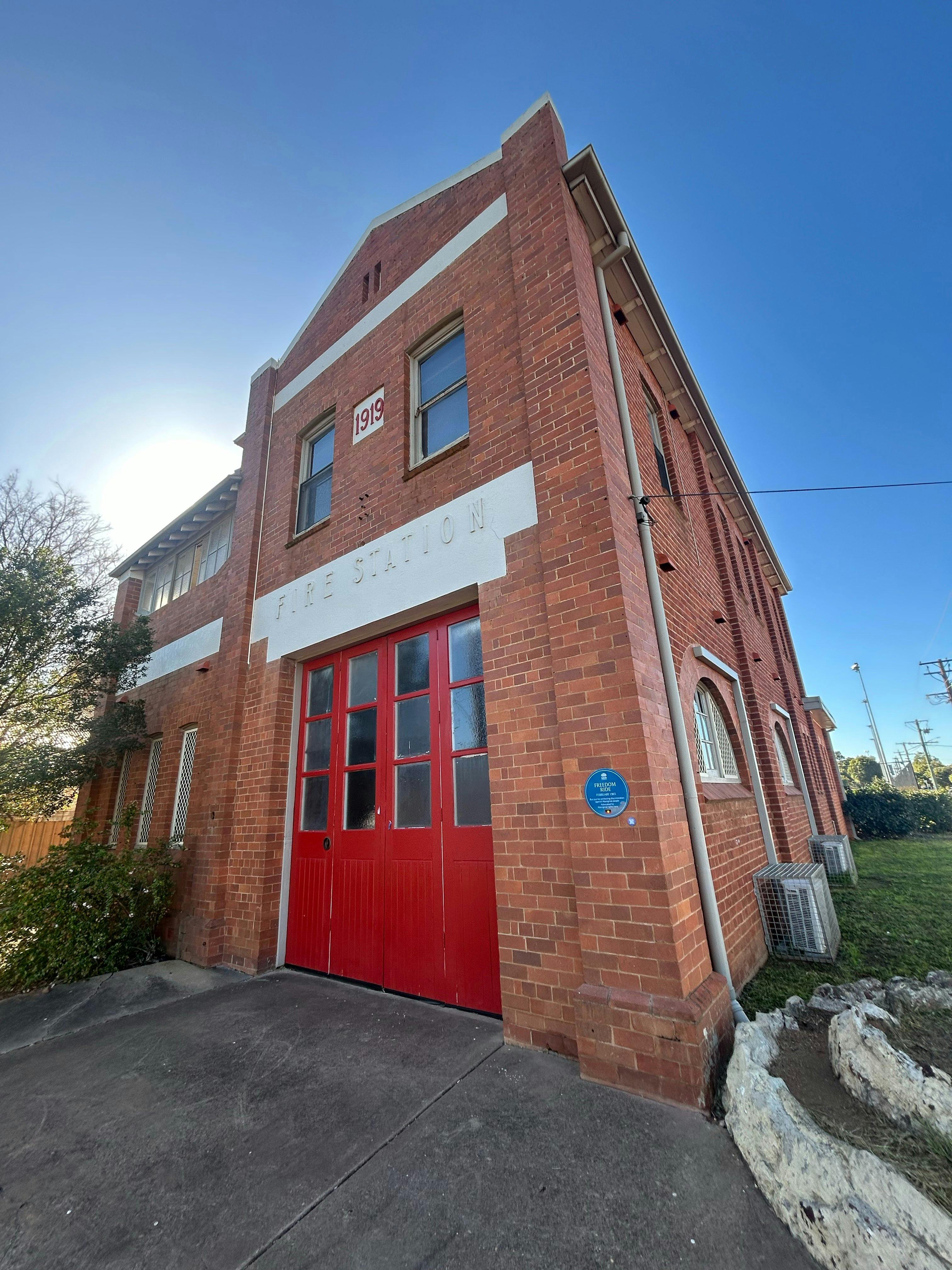 Blue plaque on 2-storey red brick building next to red doors with signs '1919' and 'Fire Station'