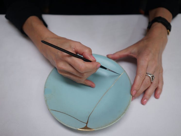 Picture shows hands holding a blue plate with a gold brush line
