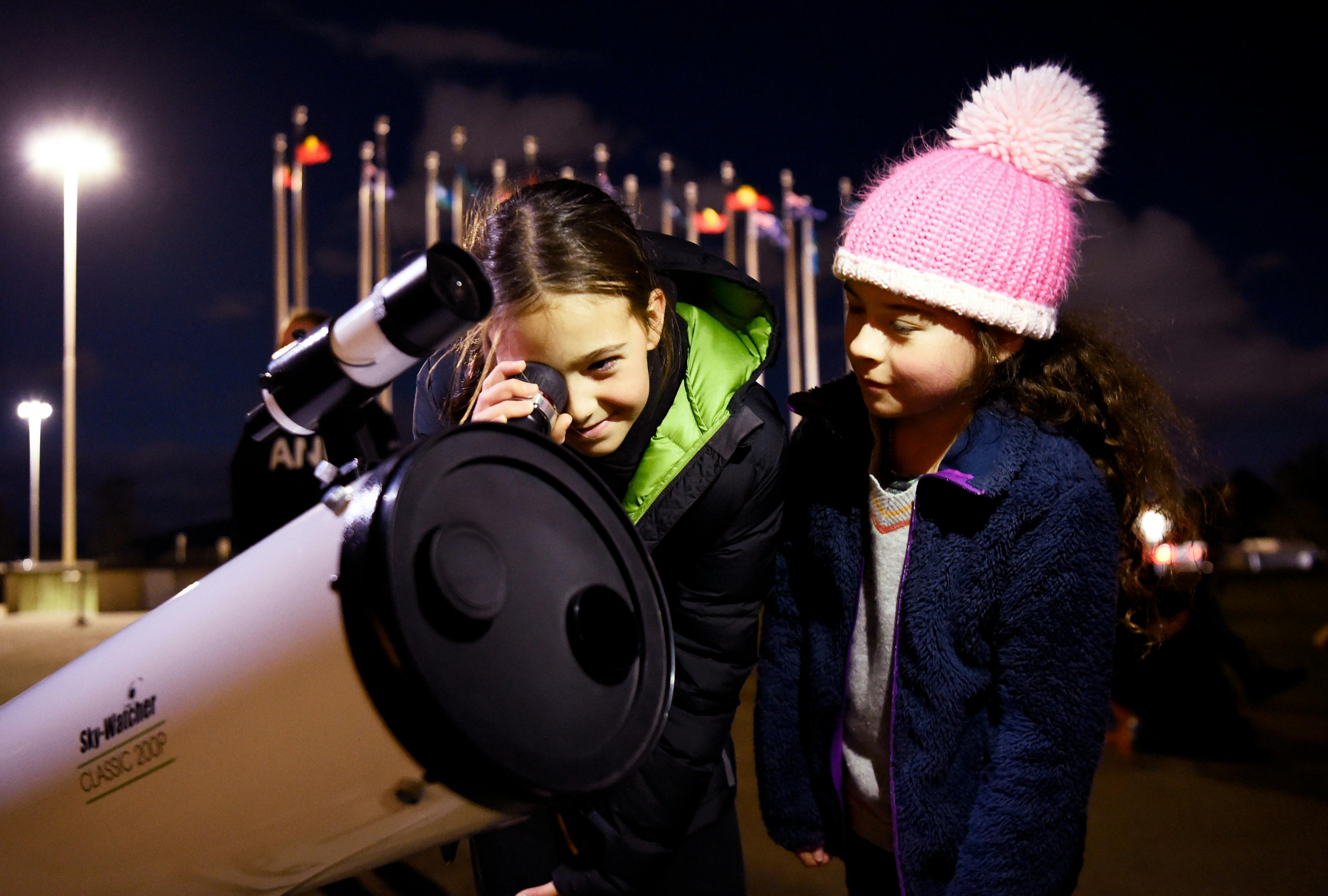 Two children stand outdoors at night using a telescope.