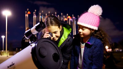 Two children stand outdoors at night using a telescope.