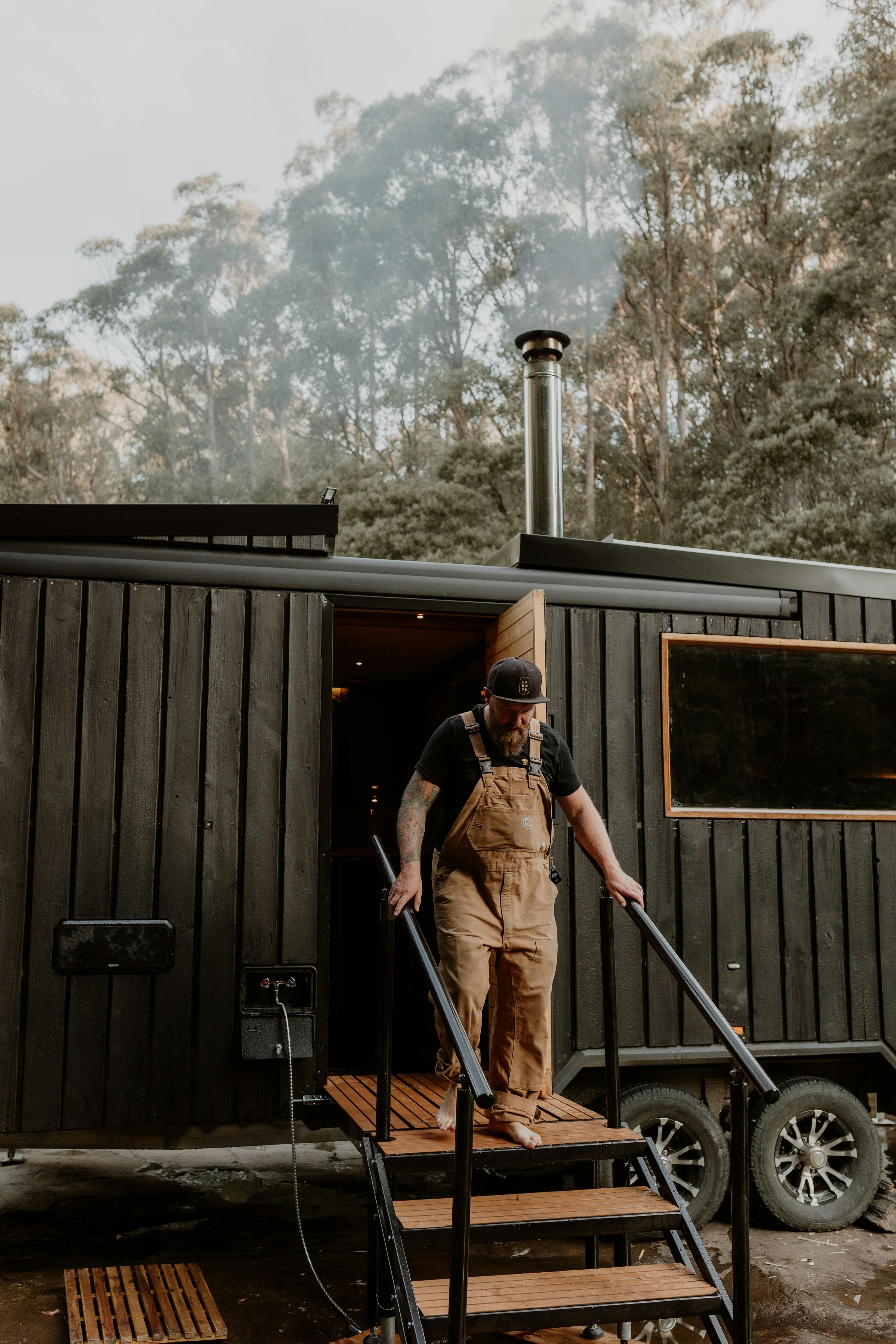 Person stepping from a wood-fired mobile sauna surrounded by native Tasmanian bushland.