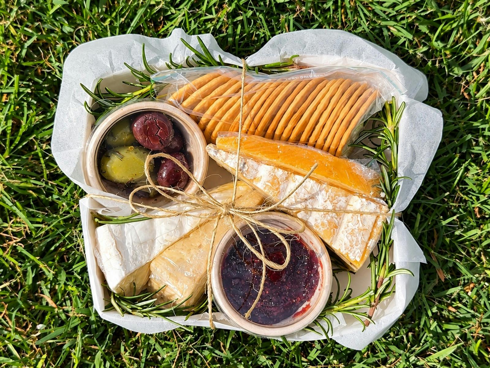 A picnic-style grazing box on green grass filled with assorted cheeses, crackers, olives & chutney.