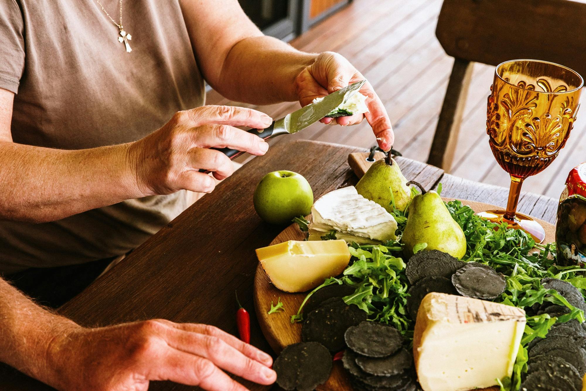 Photo of a womans hands putting cheese on a cracker with a platter of cheese, fruit, glass of wine