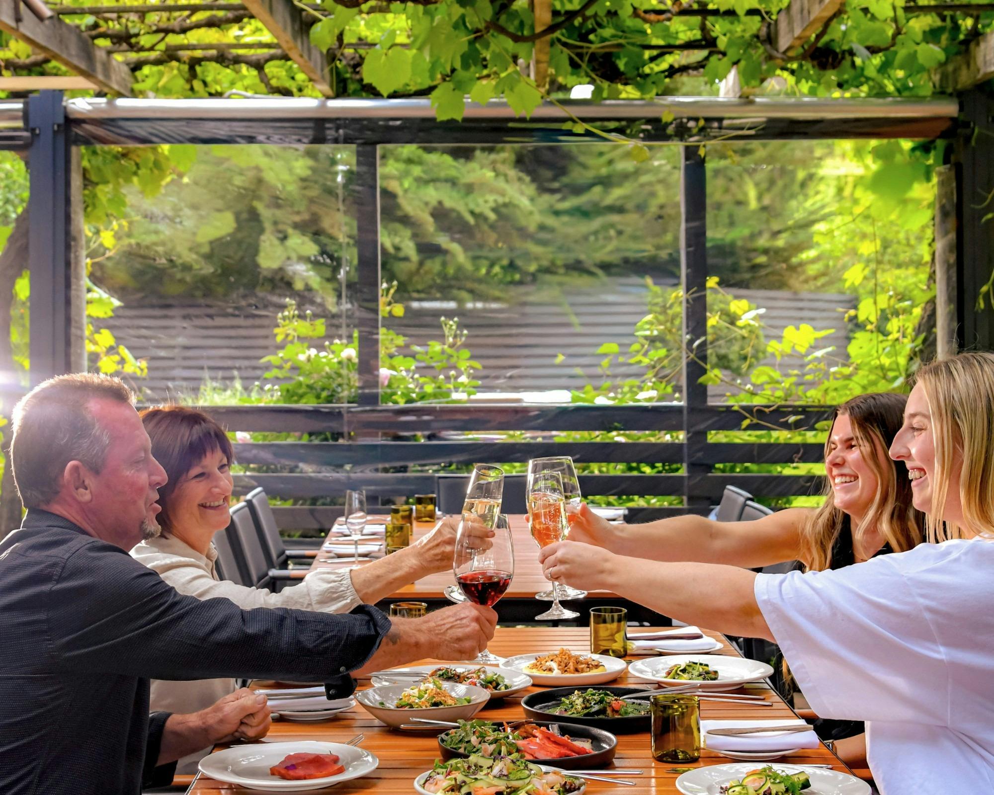 4 people clincking glasses over a table of food, beneath a vine covered arbor at The Currant Shed