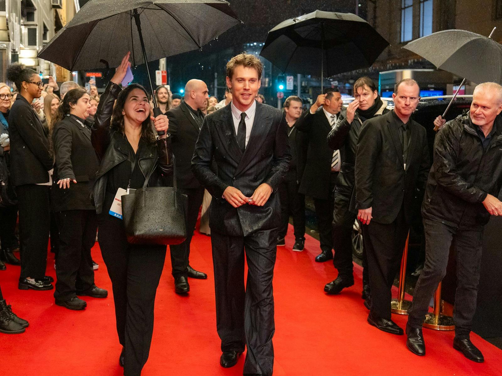 A man in a black suit walks on a busy red carpet. A woman wearing black holds an umbrella.