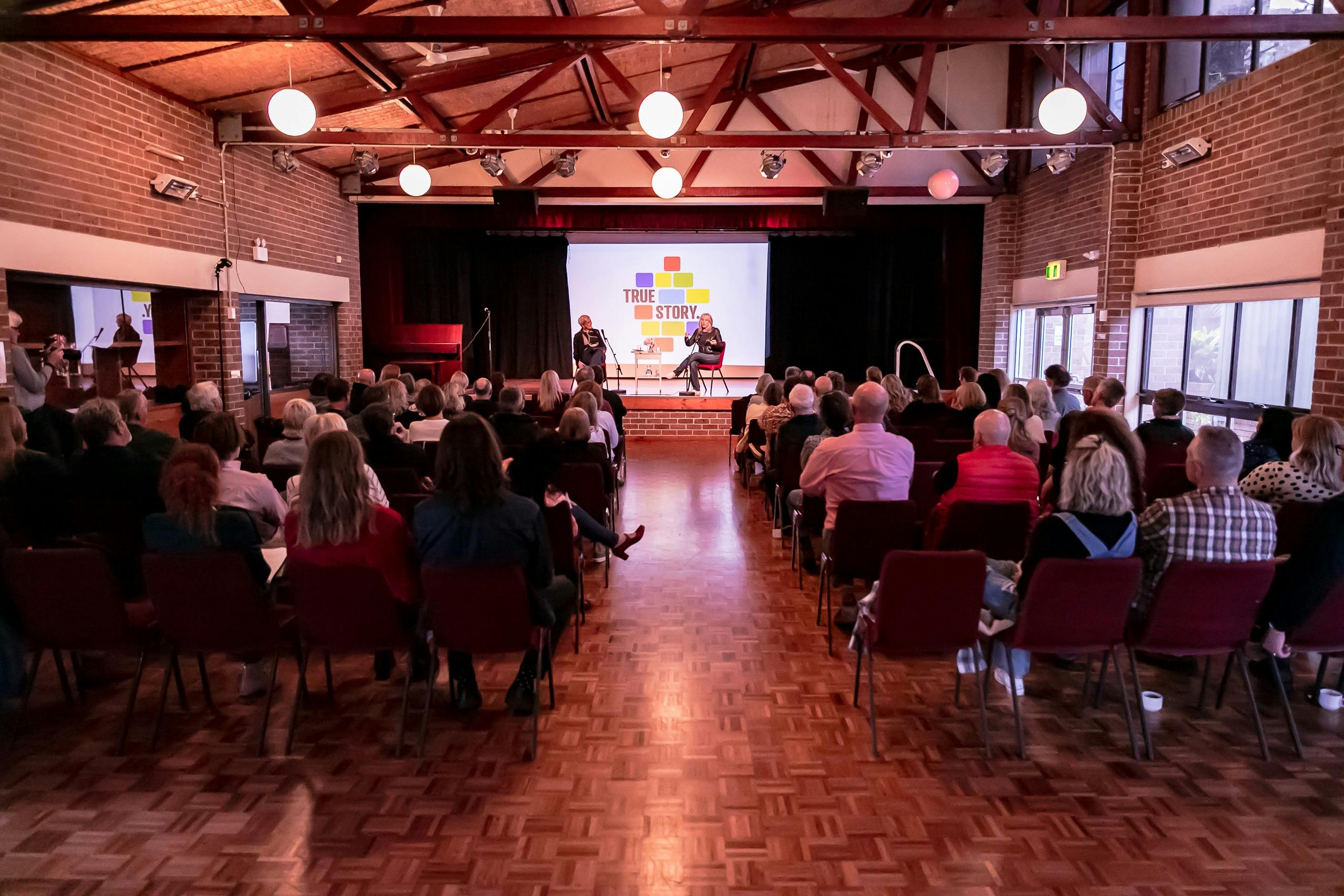 A group of people sitting in a hall, facing a stage.
