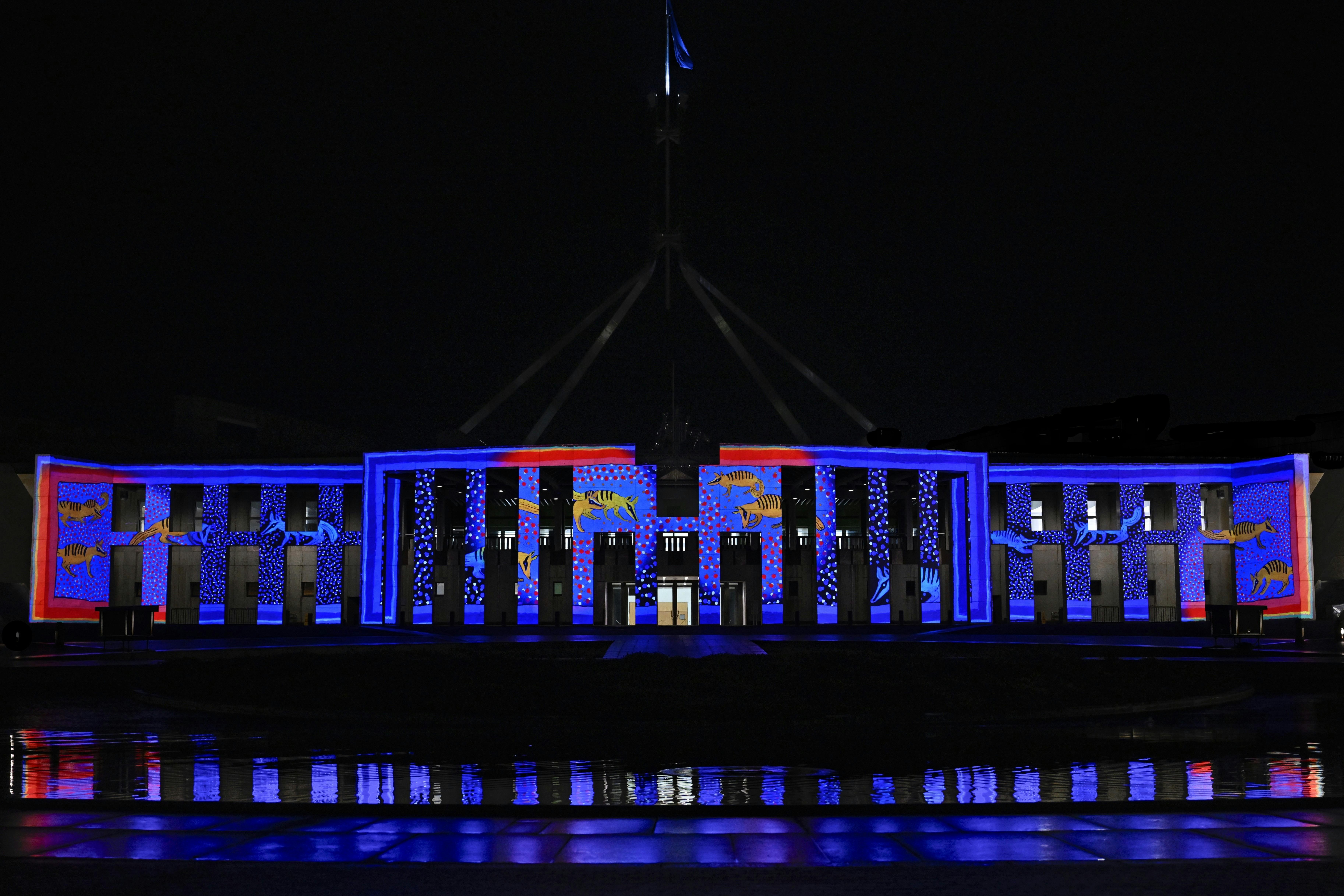 Parliament House at night, illuminated in blue & red light with projected artwork across the façade