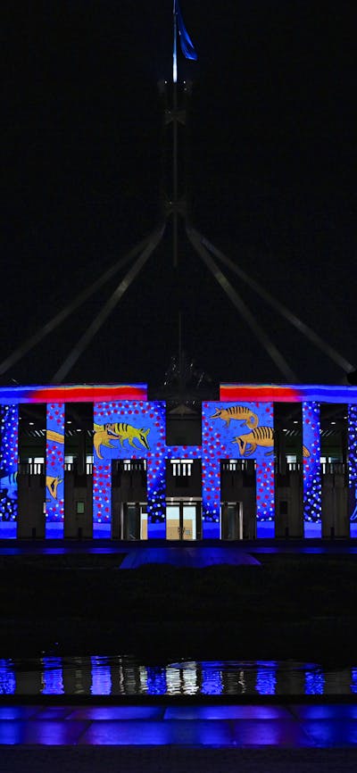 Parliament House at night, illuminated in blue & red light with projected artwork across the façade