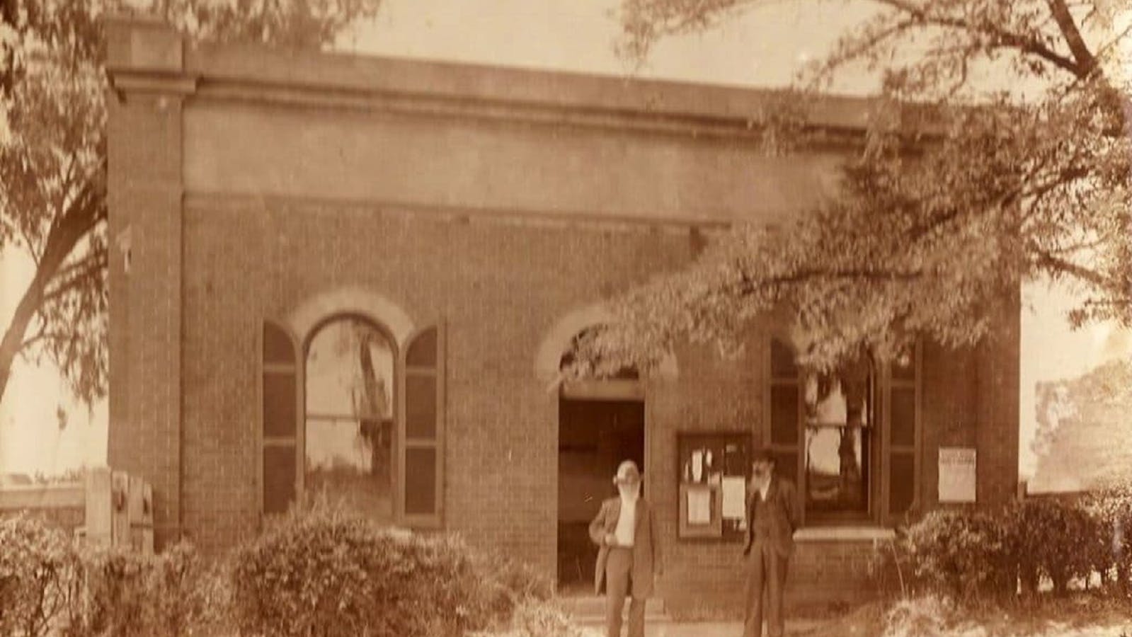 Old sepia photo of North Wangaratta Hall, with two people standing out front and trees/bushes.