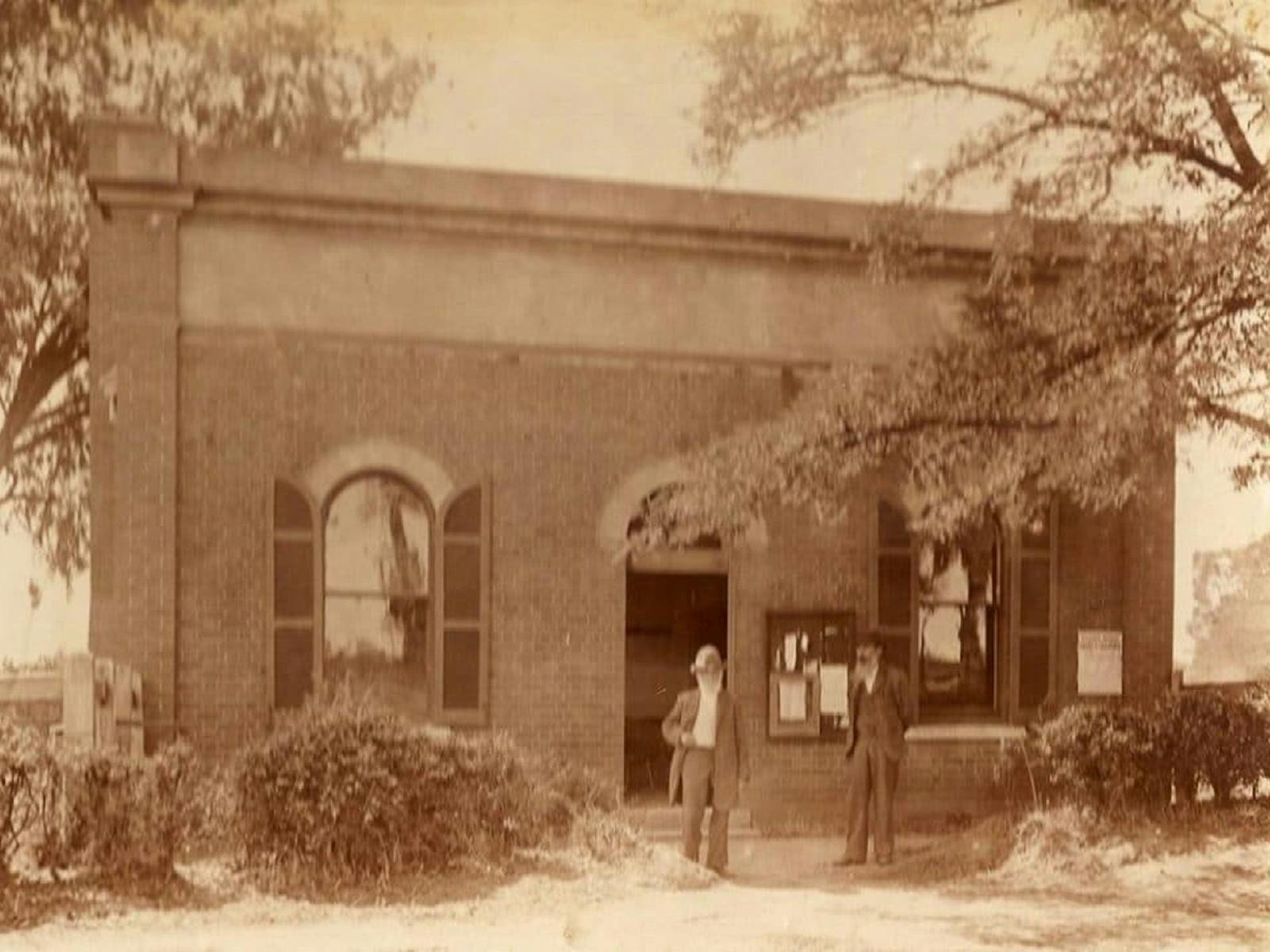 Old sepia photo of North Wangaratta Hall, with two people standing out front and trees/bushes.