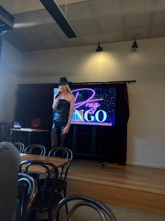 Shows a drag performer on a stage in front of a Drag Bingo sign