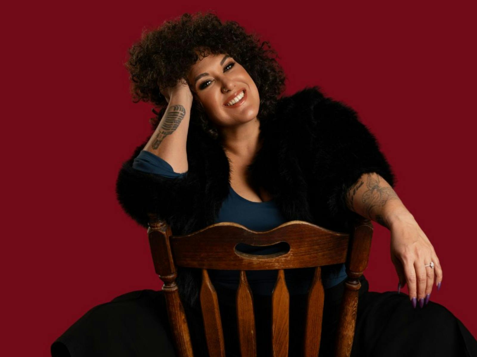 Studio portrait of a smiling woman in a black fur jacket, leaning on a wooden chair.