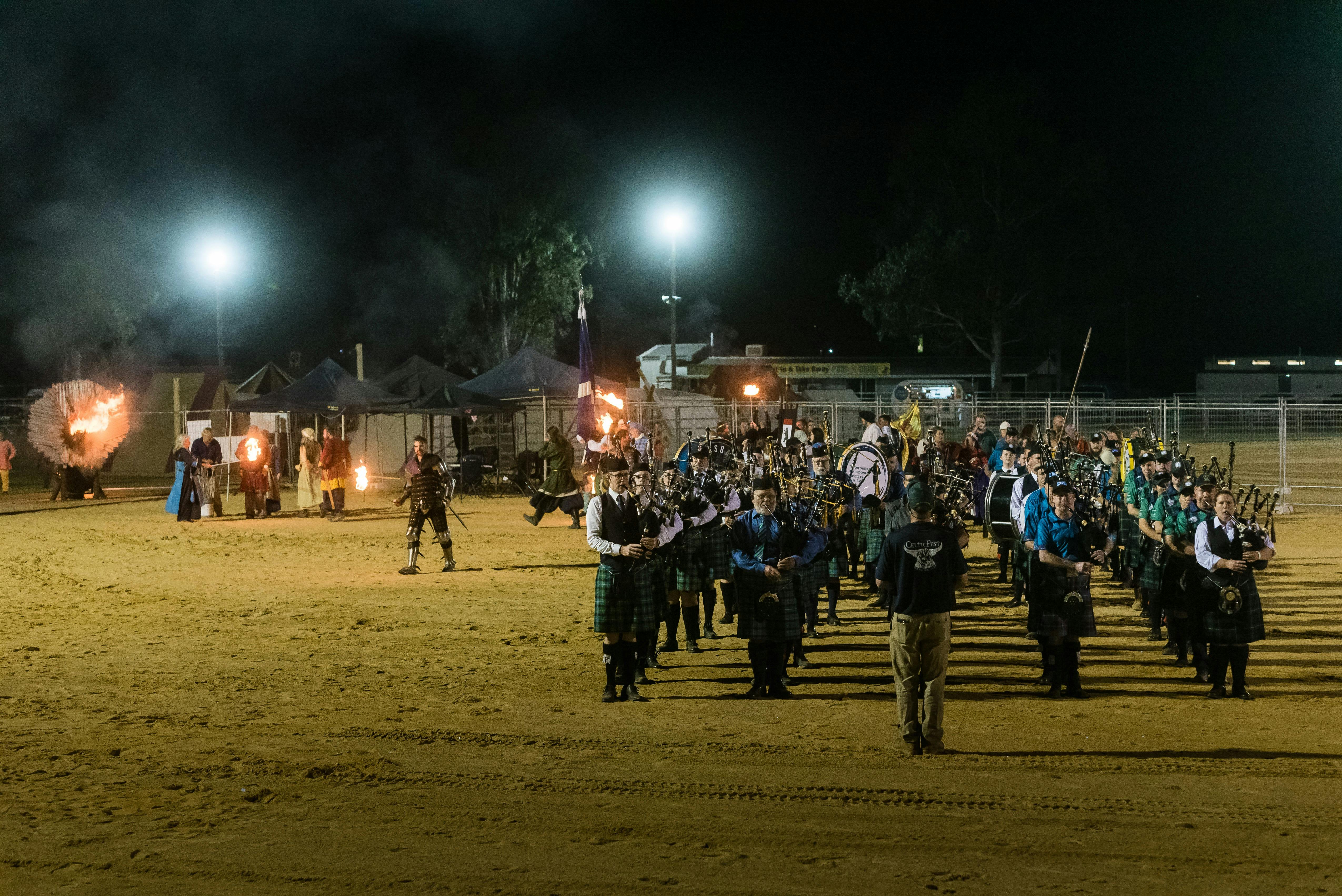 Pipers and drummers play on sandy arena, flanked by fire-breathing metal dragon and medieval knights