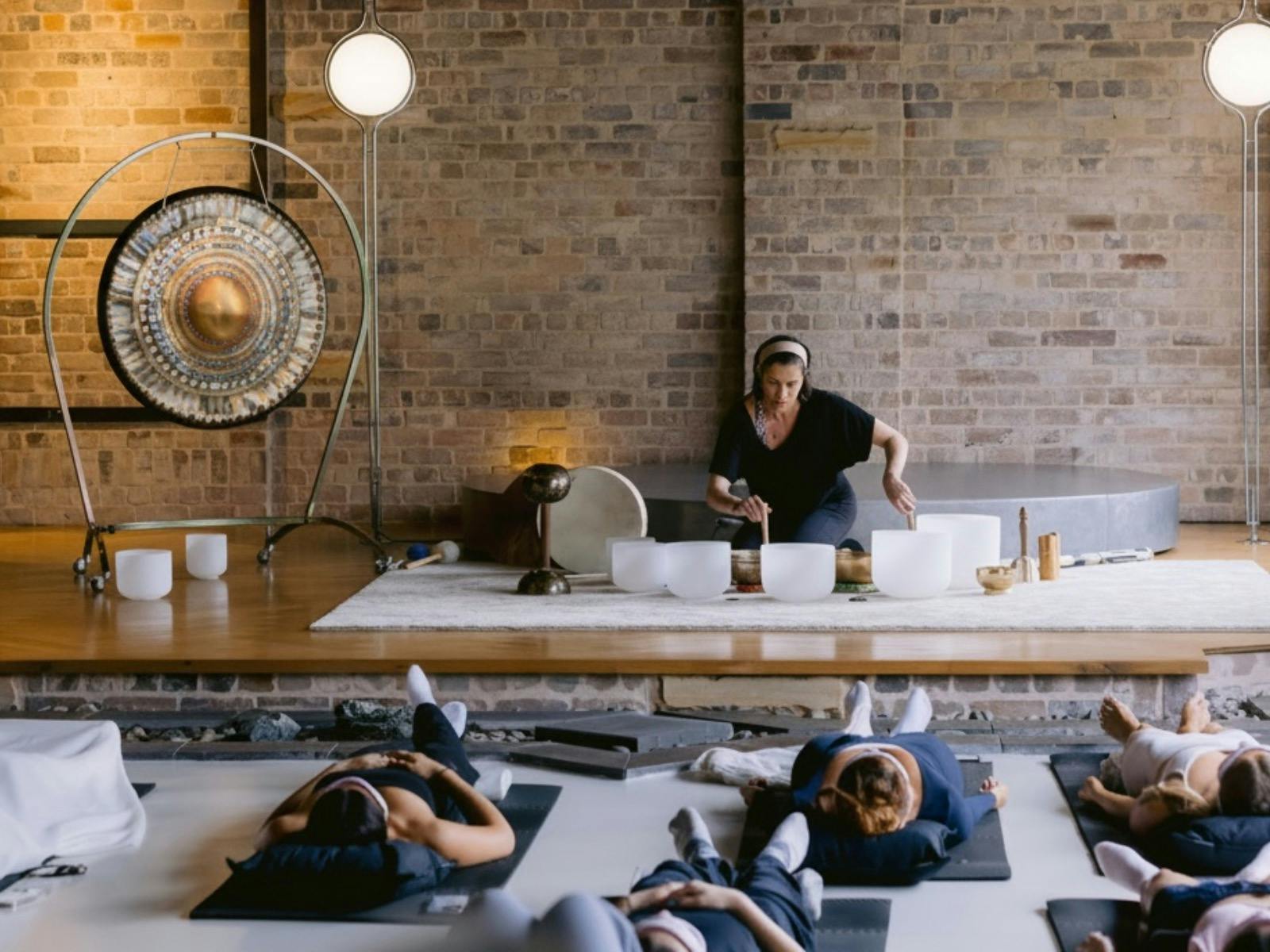 Photo of Natalie Grace playing singing bowls. People lying on yoga mats in foreground.