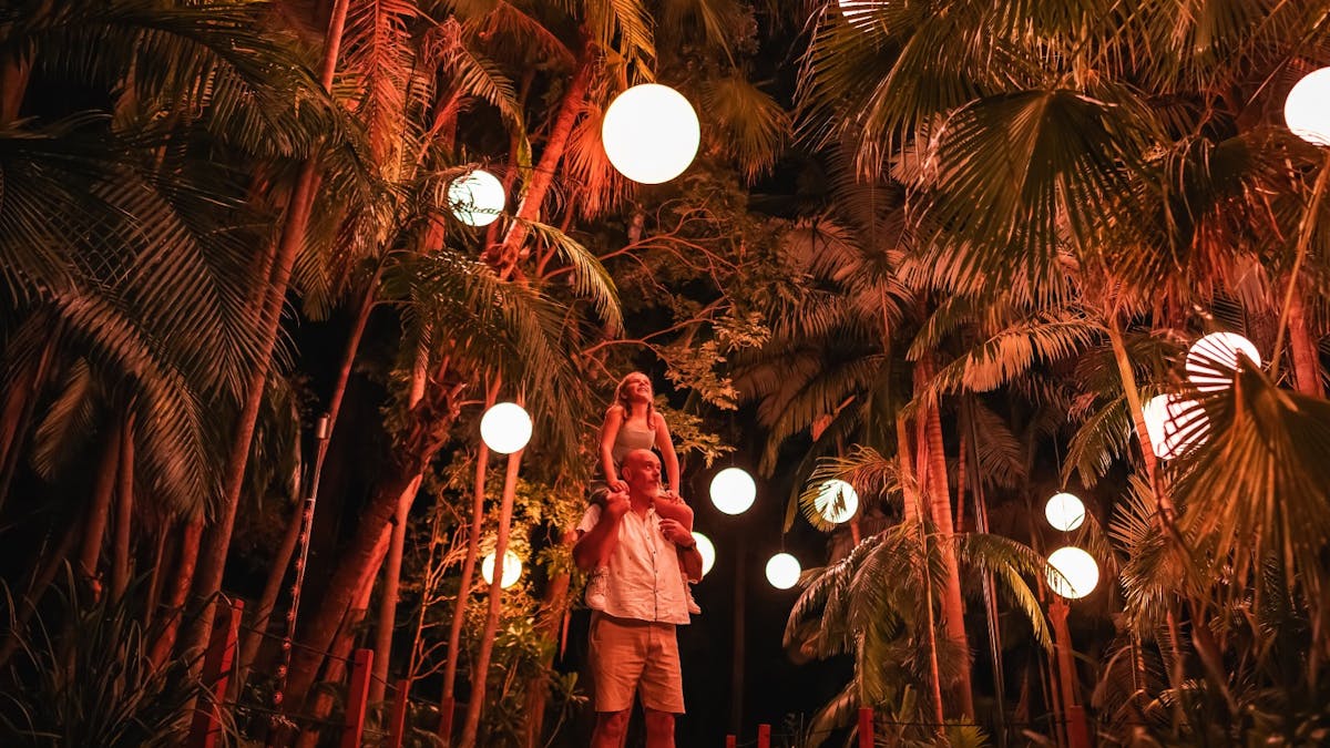 Child sitting on dad's shoulders both gazing up to illuminated balls hanging from tropical trees