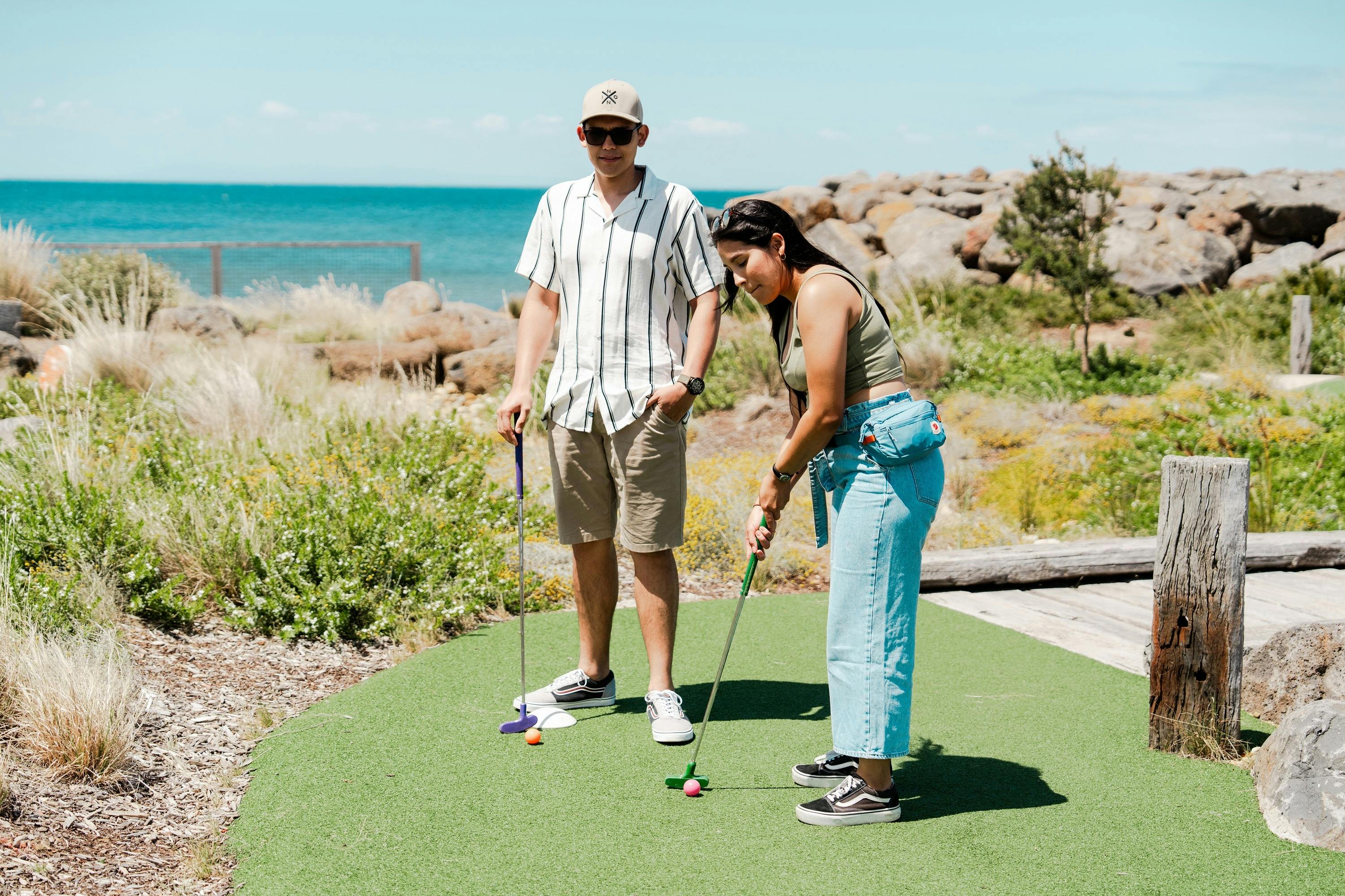 two adults playing mini golf in front of the ocean