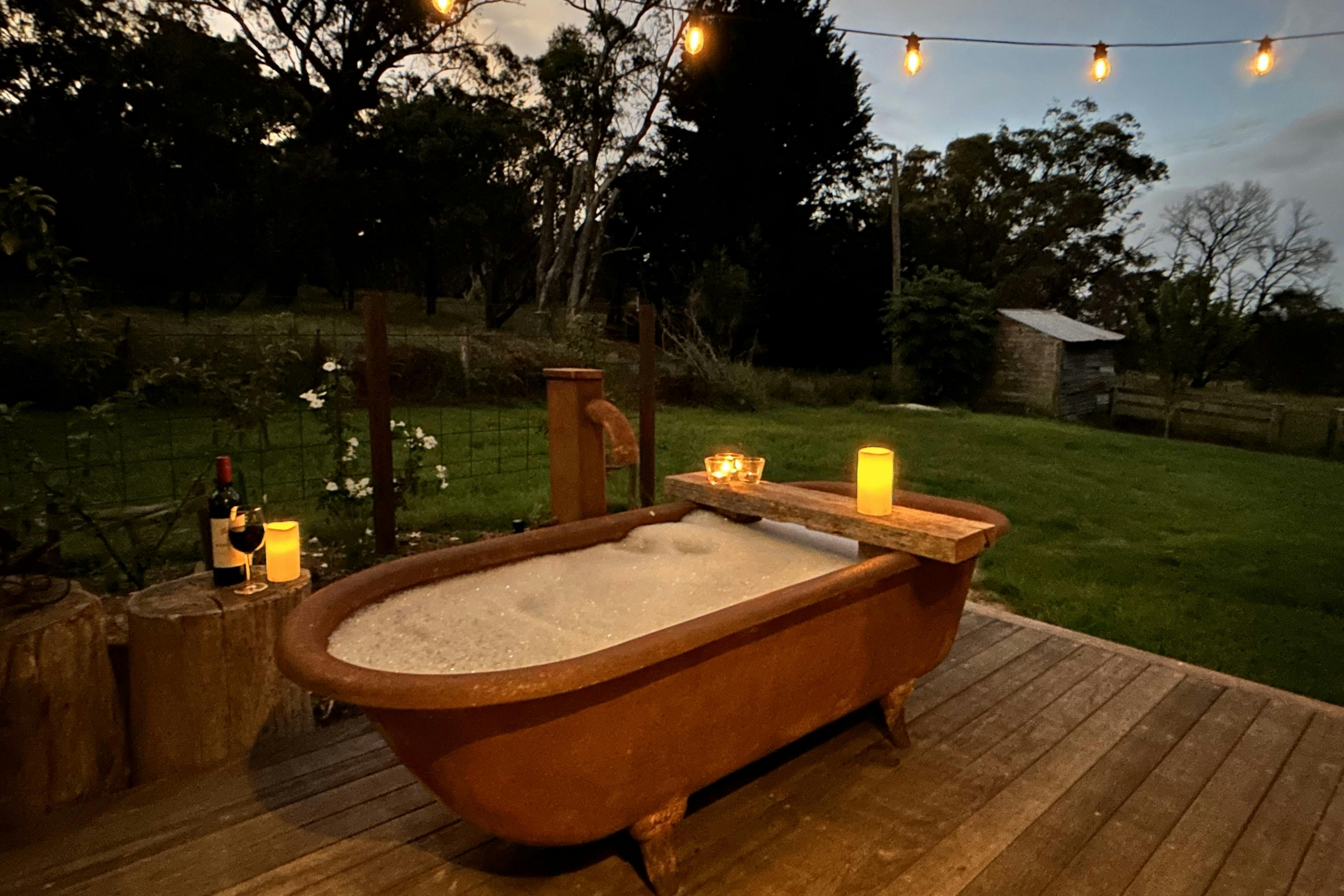 Rust-coloured bathtub at twilight on timber deck with bubblebath, candles, wine, string of lights