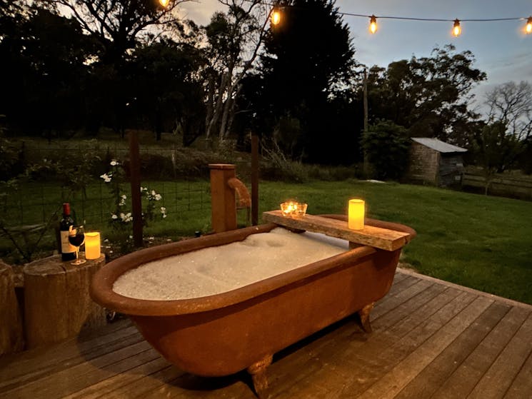 Rust-coloured bathtub at twilight on timber deck with bubblebath, candles, wine, string of lights