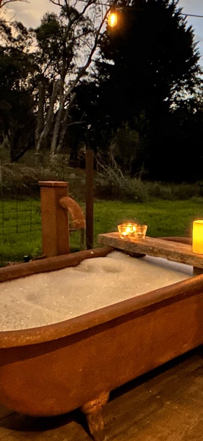 Rust-coloured bathtub at twilight on timber deck with bubblebath, candles, wine, string of lights