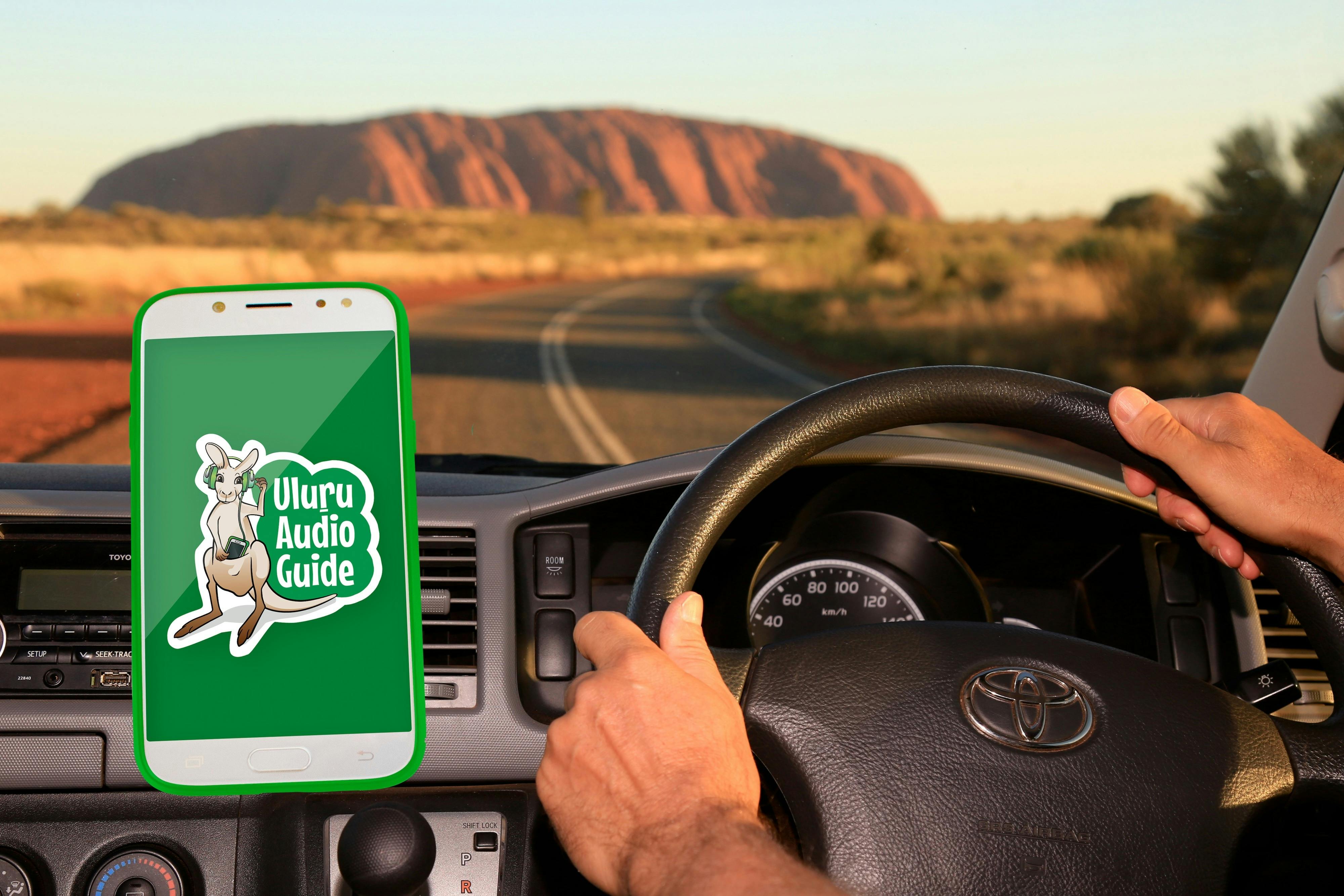 A man drives his car towards Uluru, playing the Uluru Audio Guide device through the car speaker.