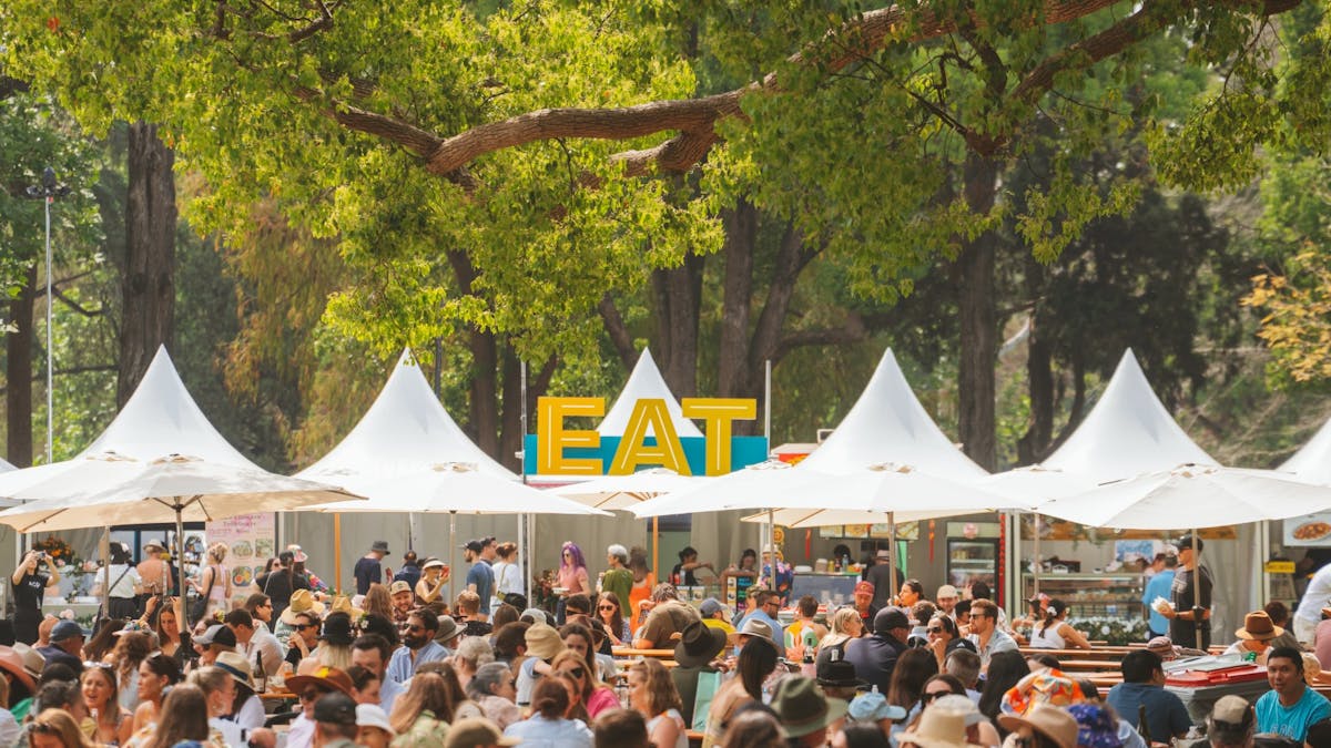 A crowd of people at an outdoor food festival set in a leafy parkland.