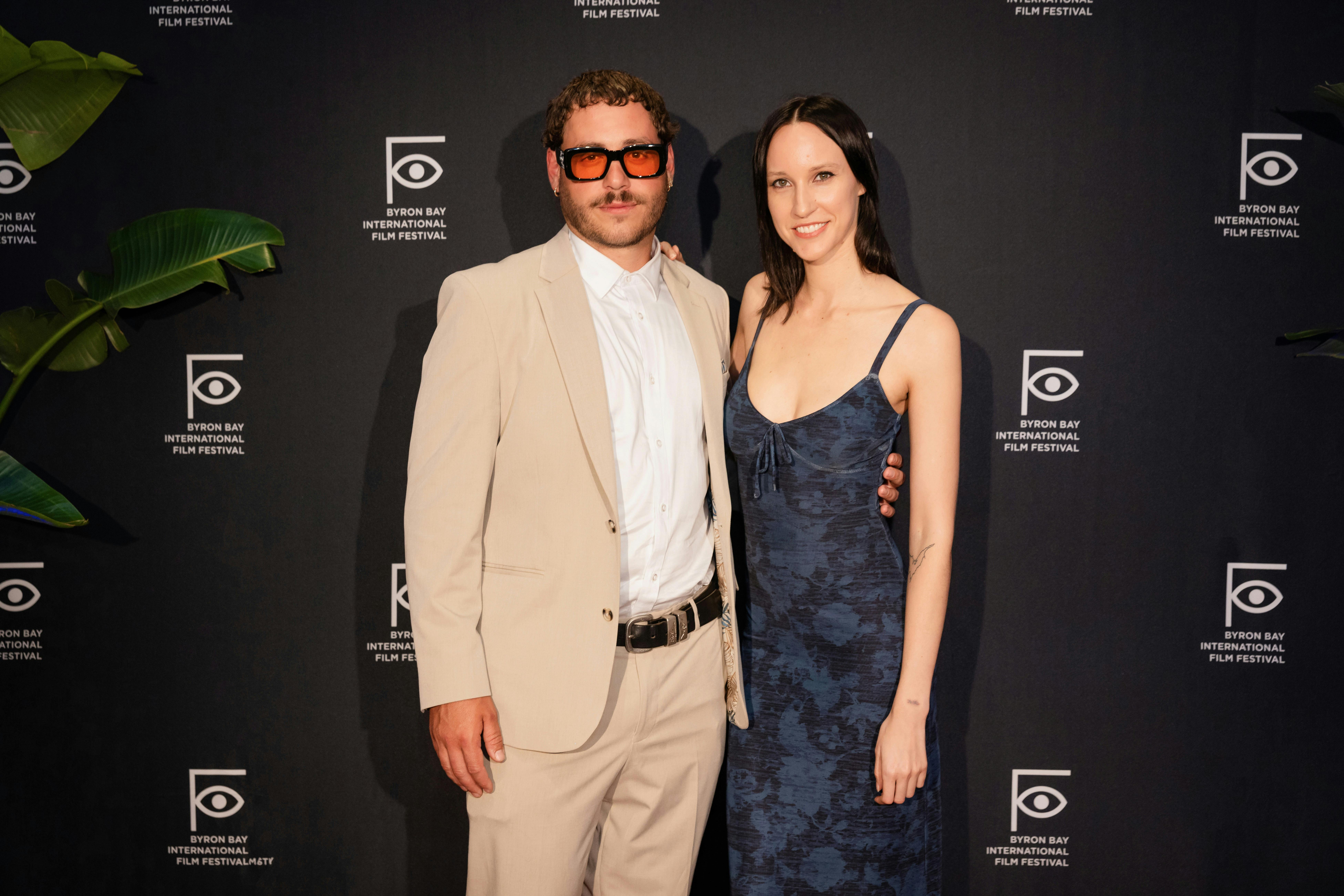 A stylish man and woman stand in front of the media wall at the red carpet opening of the festival.