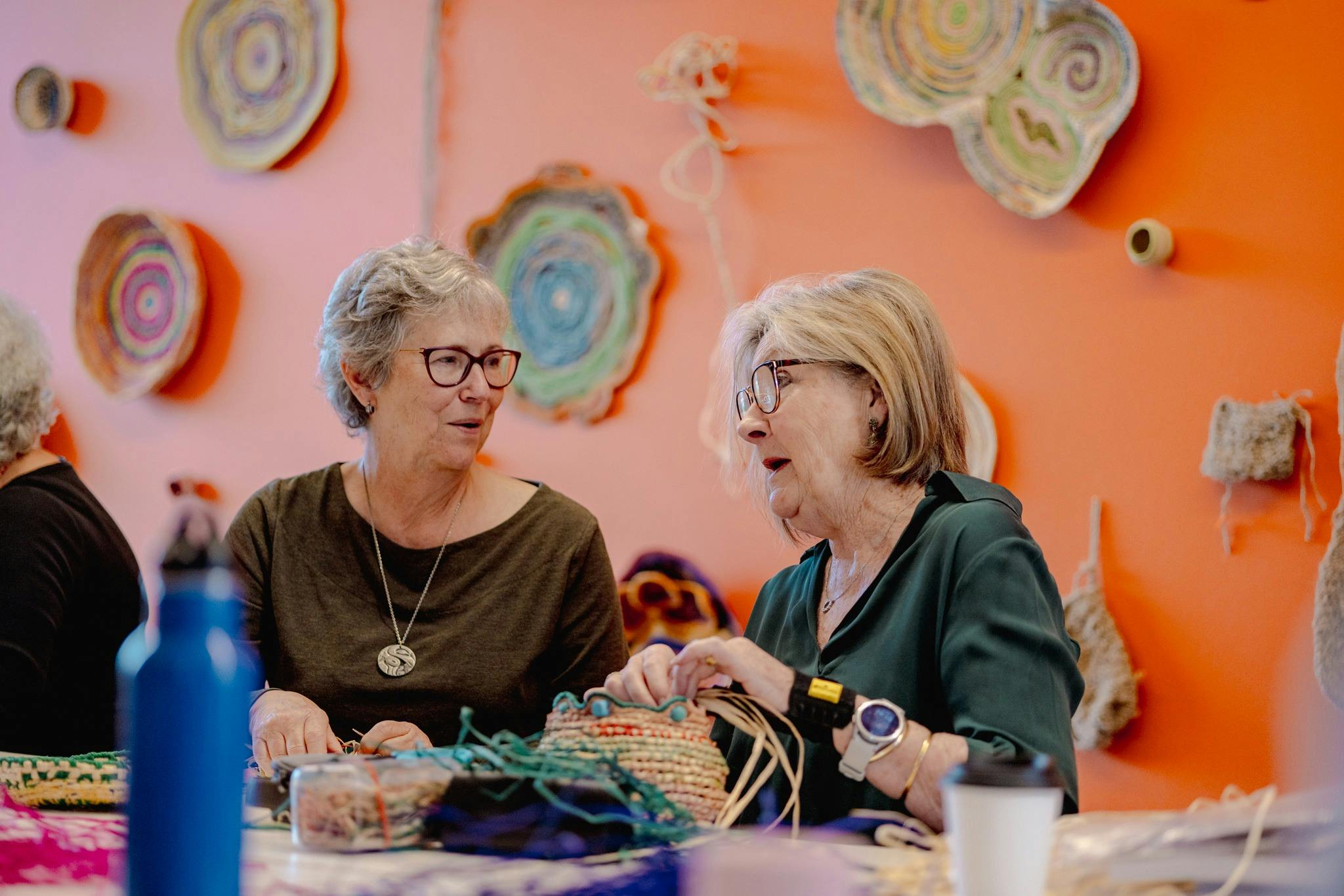 Two women having a conversation in front of an orange wall surrounded by woven baskets