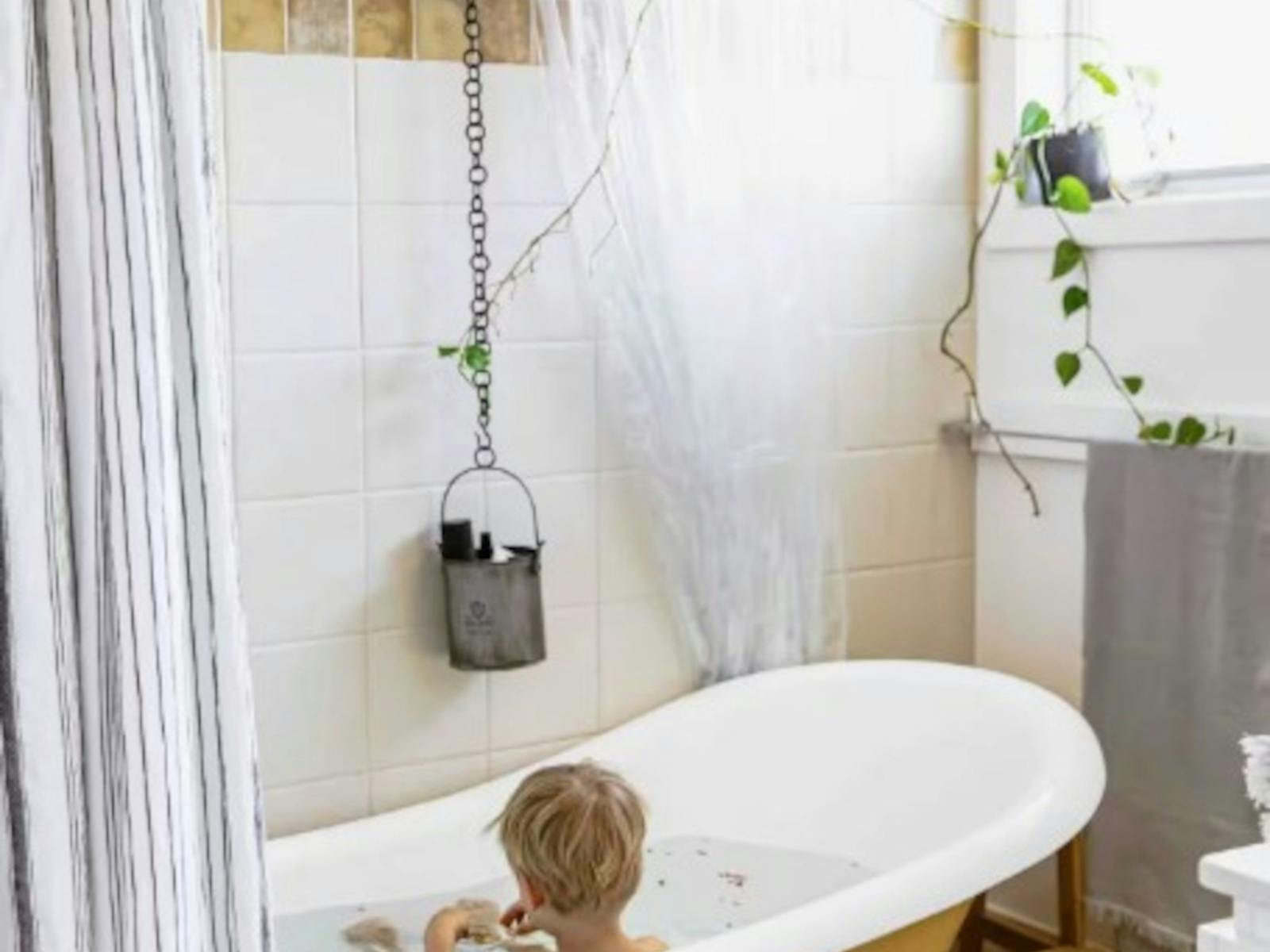 White and yellow clawfoot bath tub with child facing wall in bath. bathroom plant and white tiles.